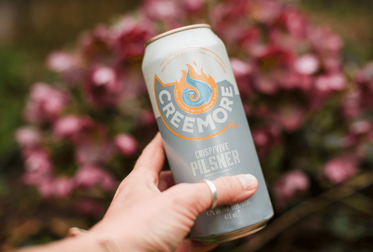hand holding creemore beer can in front of pink flowers